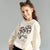A young girl with her hair in two buns wears a cream-colored sweatshirt featuring a pink and black embroidered flower design with the word 'tenderness' written below, set against a plain gray background.