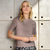 A woman wearing a short-sleeve crewneck knit top in a muted taupe-brown color, paired with a black and white patterned skirt, standing against a concrete block wall.