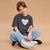 A young girl with brown hair sits cross-legged on a warm beige studio background, wearing a faded dark gray t-shirt with a white heart graphic that reads 'You are magic', light blue jeans, white socks, and black shoes, holding a light gray pouch.