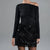 A model wearing a black off-the-shoulder long-sleeve top and a black sequined mini skirt, shown from the neck down against a solid gray studio background.