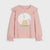 A girls' long-sleeve top in dusty pink, featuring ruffled details on the shoulders and cuffs, with a central circular graphic of a small house inside a snowy globe.