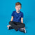 A young boy with short brown hair sits cross-legged on a solid blue studio background, wearing a royal blue athletic t-shirt with a small white logo on the left chest, black pants, and black-and-white sneakers.