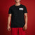 A man wearing a black short-sleeve t-shirt with a small white, red, and black chest graphic, paired with red pants, standing against a solid red background.