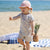 A toddler walking on a sandy beach wearing a light pink sun hat and a short-sleeve cream romper with a blue floral print. A blue and white striped towel and a pineapple are visible in the background near the ocean.
