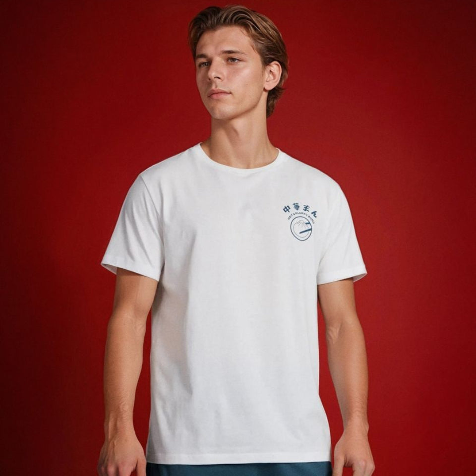 A young man modeling a white short-sleeve crewneck t-shirt with a small blue circular graphic on the left chest, paired with teal shorts, set against a solid dark red studio background.