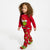 A toddler with curly brown hair wearing a red Grinch-themed pajama set with a large Grinch graphic on the top and a green-and-red all-over print on the pants, photographed against a plain white studio background.