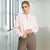 A woman wearing a light pink half-zip sweatshirt and brown plaid pants, standing in a bright, modern indoor space with large windows.