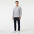 A man modeling a light gray heather crewneck sweatshirt with a small green Lacoste crocodile logo on the left chest, paired with dark navy pants and white sneakers, against a plain white studio background.