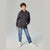 A child model wearing a dark charcoal gray hoodie with a small white graphic on the left chest, paired with blue jeans and white sneakers, standing against a light gray studio background.