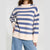 A woman wearing a cream and blue horizontally striped knit sweater with ribbed collar, cuffs, and hem, paired with light blue jeans, against a light gray studio background.