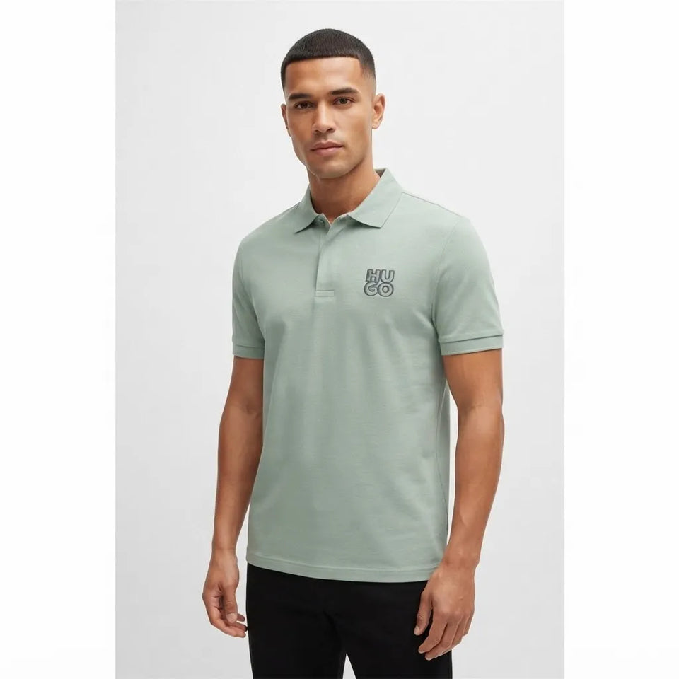 A man wearing a light sage green short-sleeve polo shirt with a small black embroidered logo on the left chest, paired with black pants, against a light gray studio background.