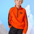 A young boy wearing a bright orange quarter-zip fleece pullover with black side panels and a small red logo patch on the chest, posed against a light blue backdrop with white mountain silhouettes.