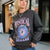 A woman wearing a dark charcoal gray sweatshirt with a colorful graphic print that reads 'STAY MAGICAL MYSTICAL DREAMS' around a blue and pink eye design, photographed outdoors in front of a storefront.