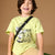 A young boy with dark hair sits smiling on a green stool, wearing a light yellow-green graphic t-shirt with a black strap across his chest and beige pants, set against a solid brown studio background.