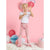 A young girl with blonde pigtails stands barefoot on a white shag rug, wearing a white short-sleeve top and pink velvet flare pants. She is posing with her hands up against a pink background decorated with pink, red, and blue balloons.