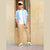 A young boy stands in a doorway wearing a light blue button-up shirt over a white t-shirt, beige cargo pants, and green and white sneakers, with a beige baseball cap.