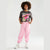 A child model wearing a dark gray graphic t-shirt with a Pusheen the cat and pink cherries, paired with light pink jogger pants and white sneakers, standing against a light gray studio background.