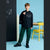 A boy modeling a black hoodie with a white graphic print, dark green sweatpants, and black sneakers, standing in a studio setting with a blue background, chalkboard wall, and yellow chair.