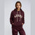 A woman modeling a maroon pullover hoodie with cream drawstrings and a cream 'ADOPE' graphic across the chest, paired with matching maroon satin track pants, against a light gray studio background.