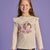 A young girl with long light brown hair stands against a solid purple studio background, wearing a beige long-sleeve t-shirt with ruffled shoulders and a sleeping hedgehog graphic, paired with dark gray leggings featuring a polka dot pattern.