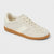 A low-top sneaker in off-white canvas with a gum rubber sole, featuring decorative ruffled stitching details around the eyelets and toe box.
