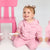 A smiling toddler girl wearing a pink hooded sweatshirt and matching pants sits on a white floor next to a pink chair, holding a white and pink toy.