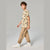 A child model wearing a beige short-sleeve t-shirt with a black graphic print and tan jogger pants with vertical black text, paired with white sneakers, set against a plain gray studio background.