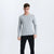 A man modeling a light gray long-sleeve V-neck knit sweater with black pants, standing against a plain white studio background.