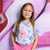 A young girl with long light brown hair and pink-framed glasses stands against a pink and purple brick wall with graffiti, wearing a light blue short-sleeve t-shirt with a pink and blue floral pattern and bright pink shorts.