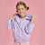 A young girl with her hair in pigtails smiles while wearing a light purple hooded zip-up jacket with small white floral prints, posed against a solid pink background.