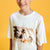 A smiling young boy with brown hair wearing a cream-colored t-shirt with a tropical palm tree graphic, set against a solid yellow background.