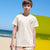A boy wearing a cream-colored t-shirt with a black graphic text print on the left chest, standing on a beach with a bright yellow wall in the background.