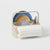 A beige plastic and metal dish caddy organizer holding a blue plate, wooden plate, glass, silver cutlery, and a roll of white paper towels, on a light gray background.