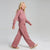 A young girl with blonde hair is wearing a matching dusty rose two-piece lounge set, consisting of a cropped long-sleeve sweatshirt and wide-leg sweatpants, walking against a light gray studio background.