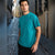 A man wearing a teal short-sleeve t-shirt with a small blue graphic on the left chest, standing on a cobblestone street.