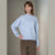 A woman modeling a light blue heathered knit sweater with a mock neck and long sleeves, paired with brown trousers, against a neutral beige studio background.