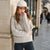 A woman wearing a chunky knit cream turtleneck sweater, a cream baseball cap, and dark blue jeans, leaning against a stone pillar on a city street.