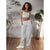 A woman modeling a light gray two-piece loungewear set consisting of a cropped tank top and matching wide-leg sweatpants, standing in a room with a wood floor and potted plants.