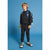 A young boy stands against a solid blue studio background wearing a black crewneck sweatshirt, black jogger pants, and black-and-white sneakers with bright green accents. He is also wearing a blue backpack with a green strap.