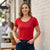 A woman wearing a bright red short-sleeve scoop-neck t-shirt and dark blue jeans, standing outdoors against a white brick wall.