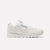 A classic Reebok sneaker in off-white leather, shown from the side against a light gray background. The shoe features white laces, a thick white midsole, and blue and red accents on the Reebok branding on the side and heel.