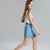 A young girl with long light brown hair is shown in profile wearing a white graphic t-shirt and a light blue denim mini skirt, set against a solid light gray studio background.