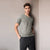 A man wearing a light sage green v-neck t-shirt and dark gray pants, standing against a neutral light gray wall.