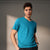 A man wearing a bright turquoise blue v-neck t-shirt and dark gray jeans, standing against a neutral gray wall with soft shadows.