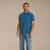 A man modeling a short-sleeve polo shirt in a solid teal blue color, paired with blue jeans and white sneakers, standing against a solid beige studio background.