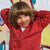A young boy with shoulder-length brown hair smiling and posing in a red zip-up hoodie against a colorful painted wall.