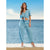 A woman stands by the ocean wearing a light blue two-piece set consisting of a short-sleeve cropped tie-front top and matching wide-leg pants, paired with white heeled sandals.