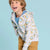 A young boy smiling and looking up, wearing a light gray zip-up hoodie with a blue and yellow dinosaur print, a blue t-shirt, and tan pants, set against a solid light blue background.
