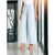A model wearing light blue wide-leg cropped pants with a pleated front, paired with a white top and beige high-heeled sandals, standing in a modern interior setting.