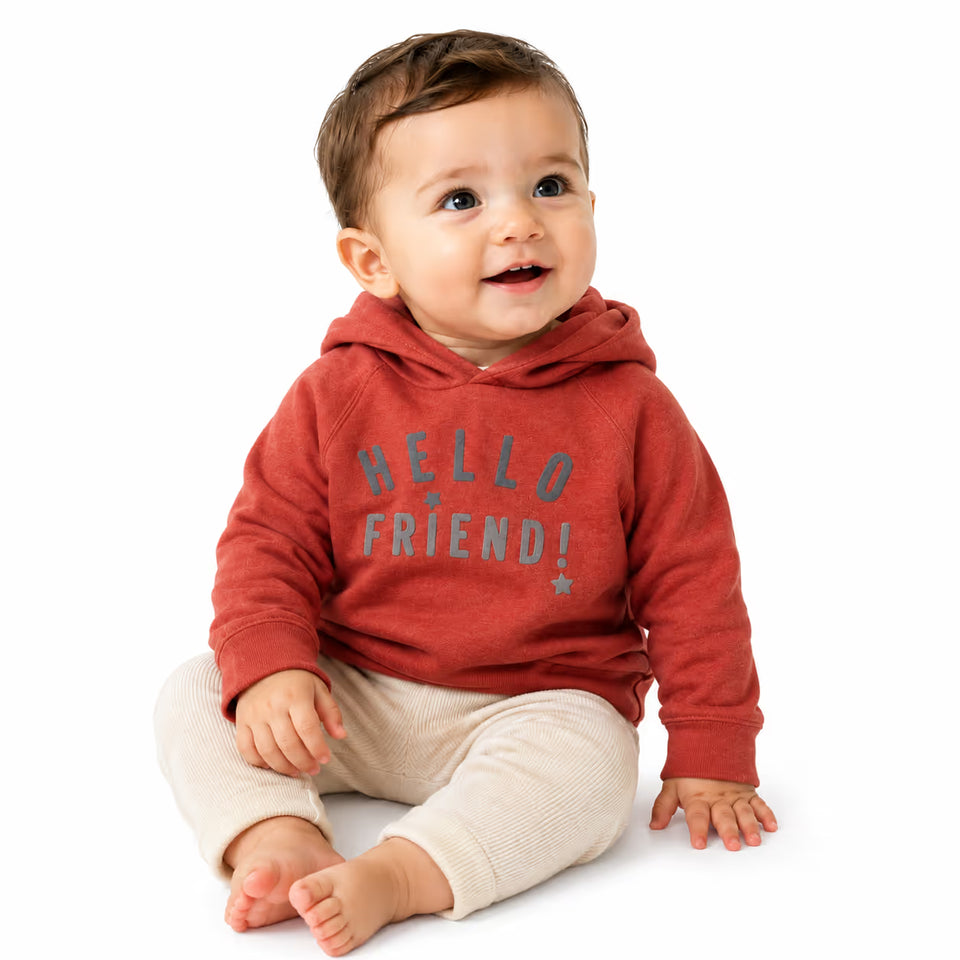 A baby sitting on a white background wearing a rust red hoodie with 'HELLO FRIEND!' printed in gray on the front, paired with cream ribbed pants.